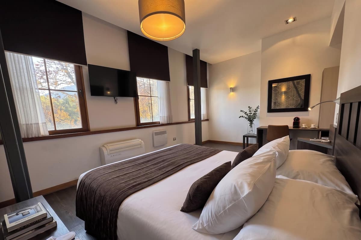 A hotel room with a bed, a flat-screen TV, a desk with a chair, a window with curtains, and a wall-mounted lamp.