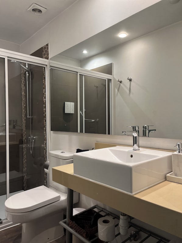 Un baño moderno con ducha envuelta en vidrio, inodoro blanco, lavamanos grande con espejo y toallero.