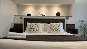 Un dormitorio moderno con una cama grande, almohadas decorativas, una mesita de noche y una repisa en la pared con adornos.