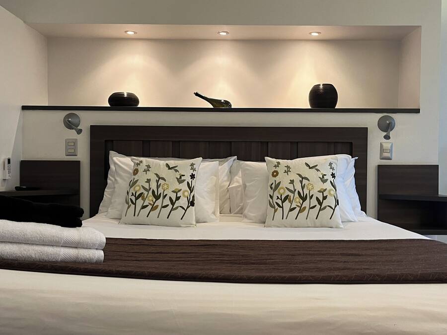 A modern bedroom with a large bed, decorative pillows, a bedside table, a nightstand, and a wall-mounted shelf with decorative items.