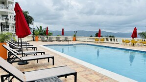 En swimmingpool med liggestole og røde parasoller, omgivet af en fliseterrasse og en hvid bygning med balkoner.