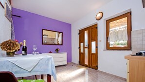 Una habitación con pared morada, una puerta de madera, una ventana con cortina, una mesa con una botella y vasos, y un aparador blanco.