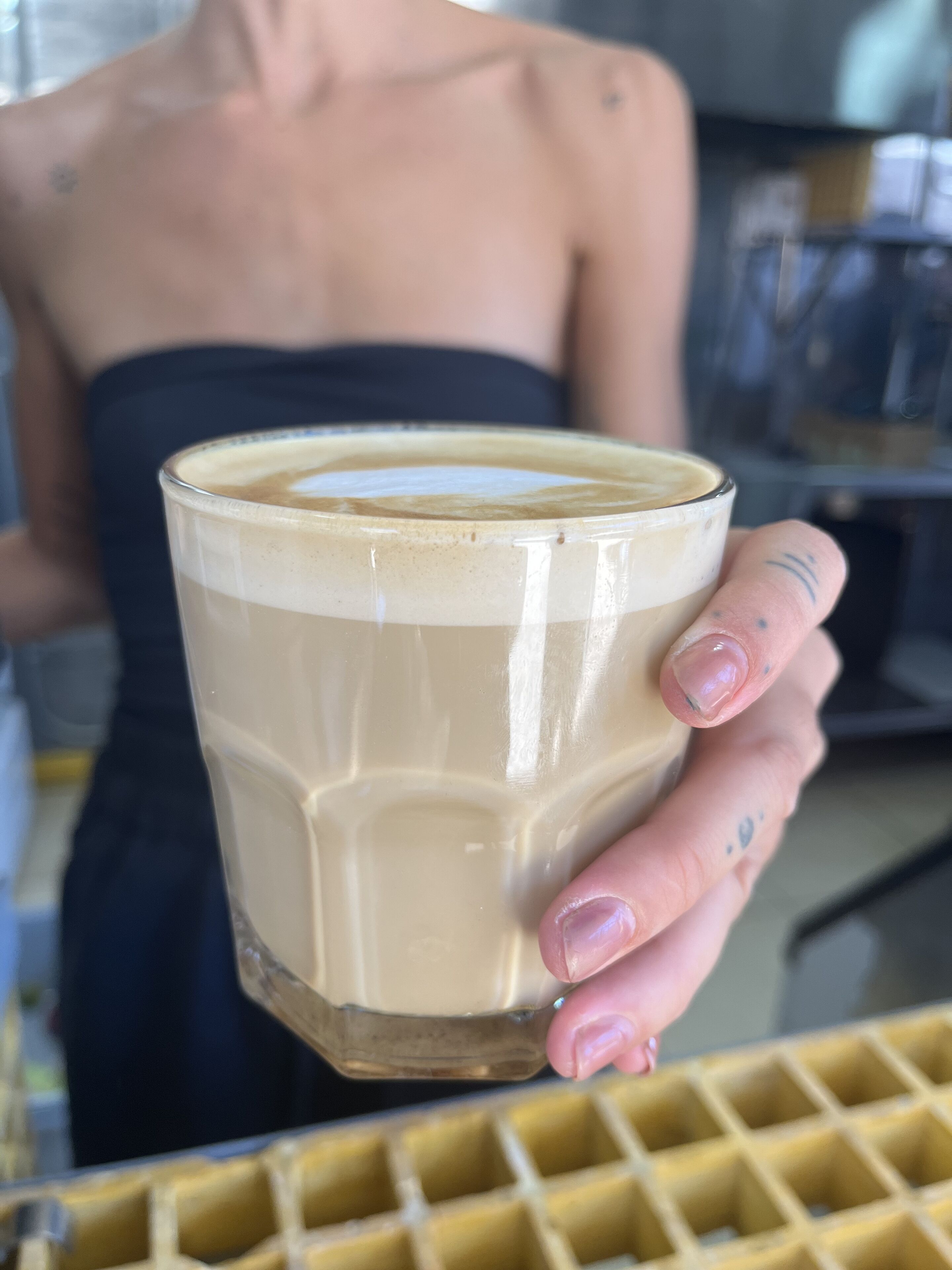 Una persona sostiene una taza de café con crema, vestida con una blusa negra y un tatuaje en la mano.