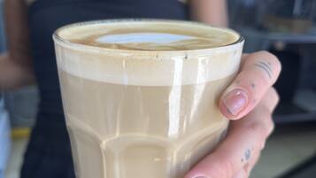 Una persona sostiene una taza de café con crema, vestida con una blusa negra y un tatuaje en la mano.