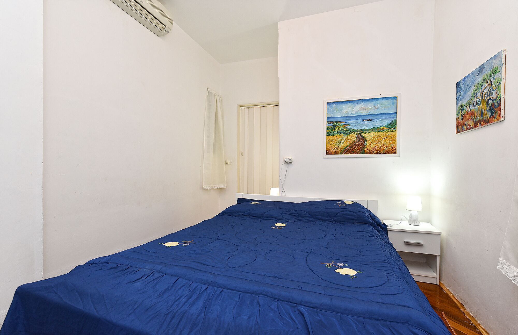 A bedroom with a single bed, a bedside table, a nightstand, and two paintings on the wall.