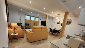 A modern living room with a sofa, a small round table, and a TV. There is a dining area with a table and chairs. The room has wooden flooring and a kitchen area with a sink and countertop.