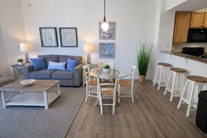 Uma sala de estar moderna com um sofá cinzento, uma mesa de centro e um espaço de jantar com uma mesa redonda e cadeiras brancas. Há quadros nas paredes, um lustre pendente e um vaso de plantas.