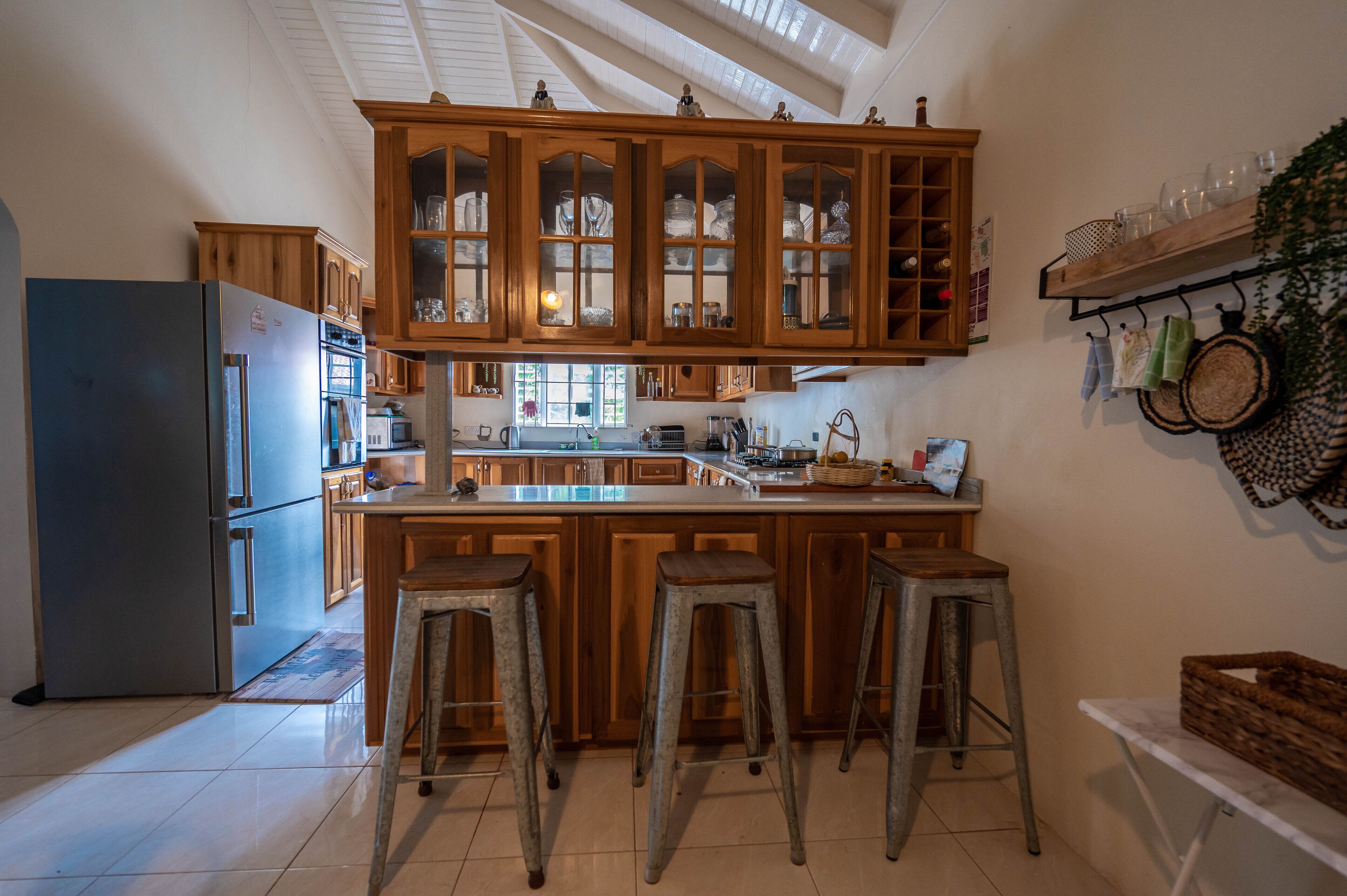 A kitchen with wooden cabinets, a central island with stools, a refrigerator, and a hanging shelf with kitchen utensils.