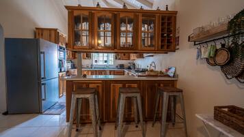 A kitchen with wooden cabinets, a central island with stools, a refrigerator, and a hanging shelf with kitchen utensils.