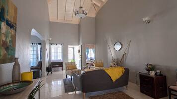 A spacious living room with a high ceiling, a grey sofa, a coffee table, and a dining area with a yellow bowl and a vase.