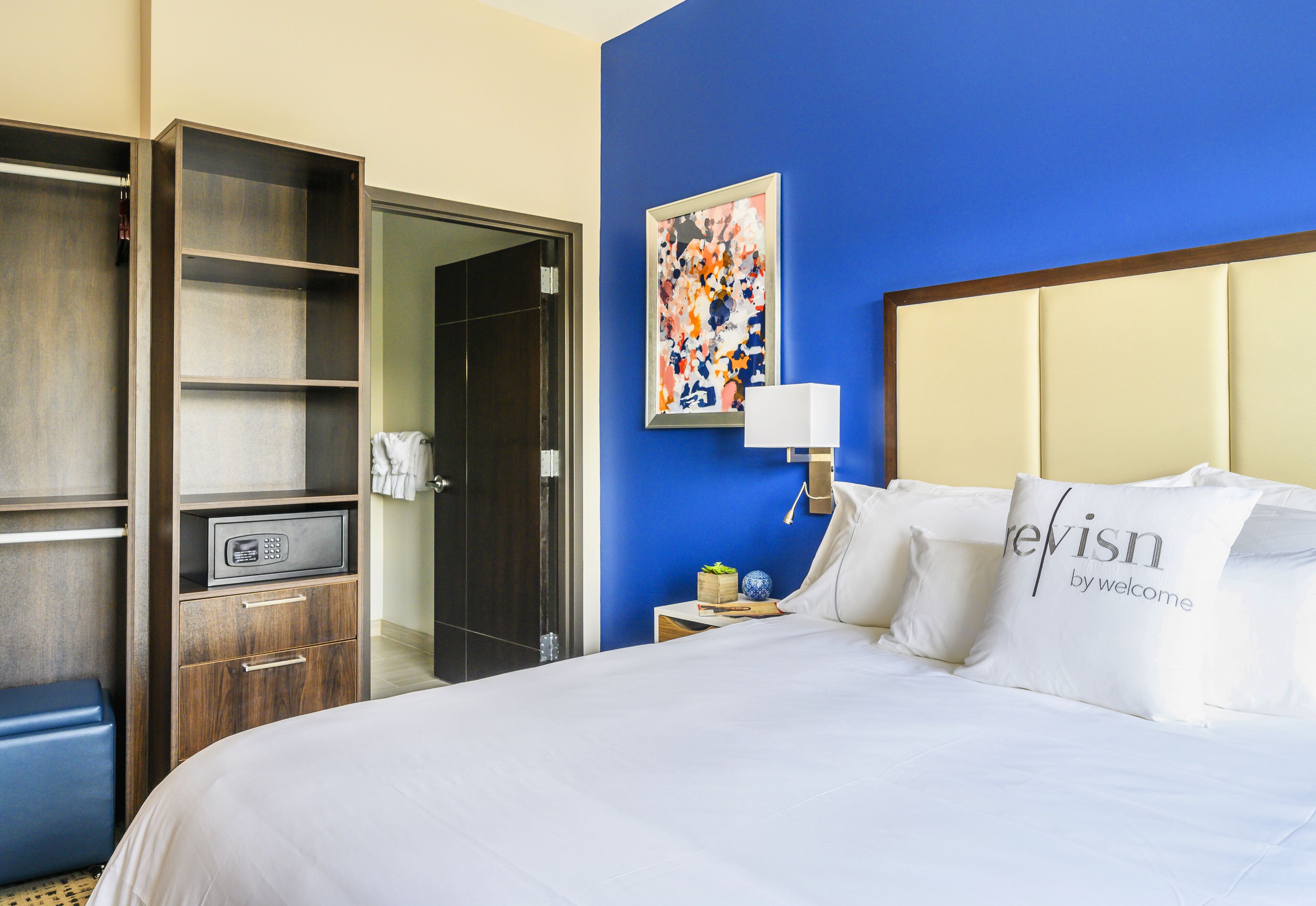 A hotel room with a bed, a wardrobe, a desk, a lamp, and a painting on the wall.