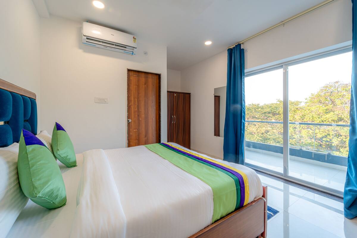 A hotel room with a large bed, a window with a view of greenery, an air conditioning unit, and a wooden wardrobe.