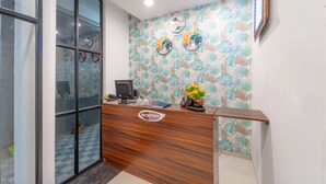 Un mostrador de recepción con detalles de madera, un letrero que dice 'Reception', una computadora y objetos decorativos, incluyendo un jarrón con flores.