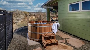 A wooden hot tub with a towel, a stone bench, and a bottle of wine on a patio with a green building in the background. - Peaceful creek-country escape that feels almost magical, with a private trail to Oak Creek and a cedar hot tub off the master bath. Very private, yet 10 min to wineries and 25 min to Sedona. Usually 10-15 degrees cooler than Phoenix. (Cornville)