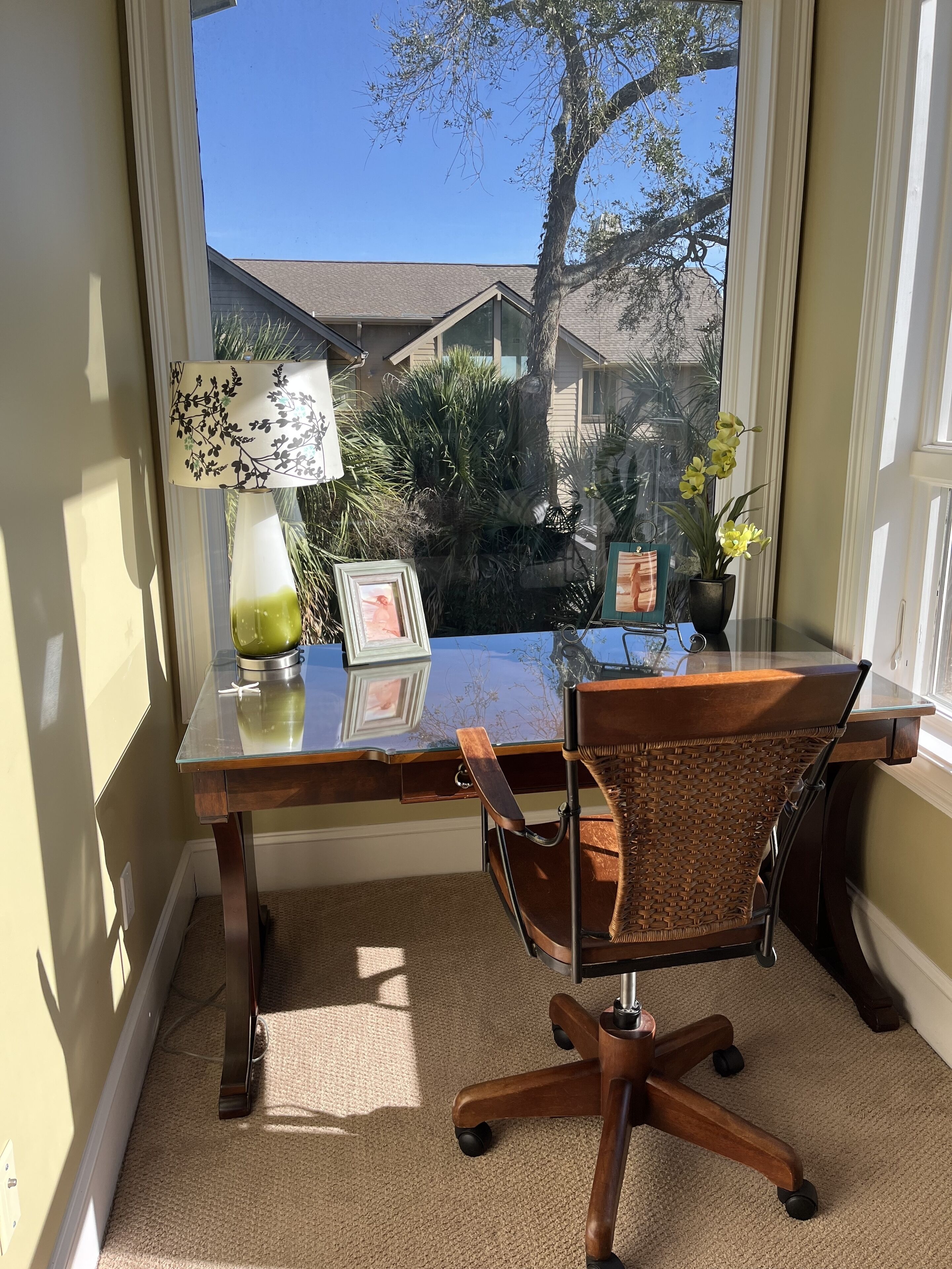 A home office with a glass desk, wicker chair, and a view of a house and trees through a window.