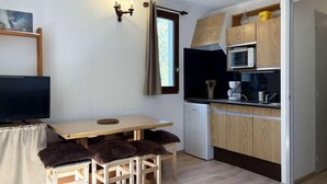 A compact kitchen with a microwave, coffee maker, and wooden cabinets. There is a dining table with two chairs and a TV on a stand.