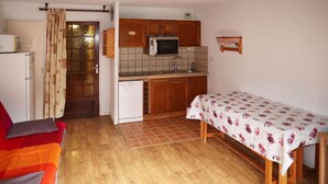 Une cuisine compacte avec des meubles en bois, un four à micro-ondes et un évier. On y trouve une petite table à manger recouverte d’une nappe à motifs floraux. Un canapé rouge et un réfrigérateur blanc sont également visibles.