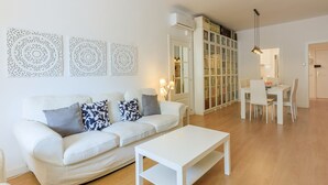 A modern living room with a white sofa, a white coffee table, and a dining area with a white table and chairs. There are decorative wall art pieces and an air conditioning unit visible. - Apartment with private terrace, 3 bedrooms, Eixample (Barcelona)