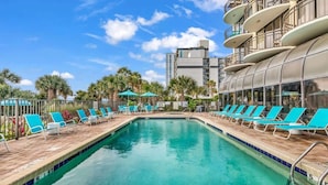 A swimming pool with lounge chairs and umbrellas, surrounded by palm trees and a modern building with balconies. - Oceanfront Penthouse, Amazing Views, Walk to Conv Center, Attractions & Dining (Myrtle Beach)