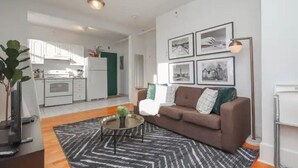 A modern living room with a brown sofa, a coffee table, and a black and white rug. There is a kitchen in the background with white cabinets and a green accent wall.