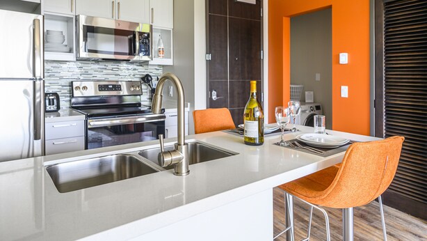 A modern kitchen with stainless steel appliances, a large island with a sink, and orange chairs. - Revisn Executive Downtown Raleigh Apartment (Raleigh)