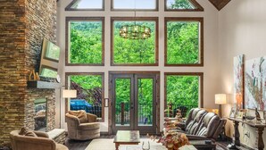 A spacious room with a high ceiling, large windows, a stone wall, and a dining area set up with plates and glasses. - Luxury Mountain Lodge • 4BR • Pool Table + PS5 • Fireplace • Views (McGaheysville)