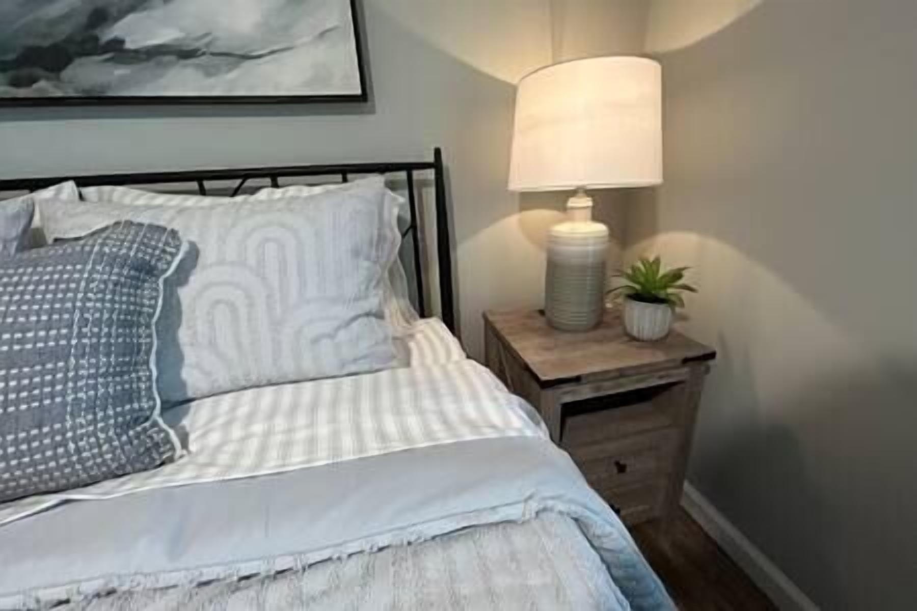 Een net opgemaakt bed met patroonbeddengoed, een bedlampje, een kleine potplant en een ingelijst beeld aan de muur.