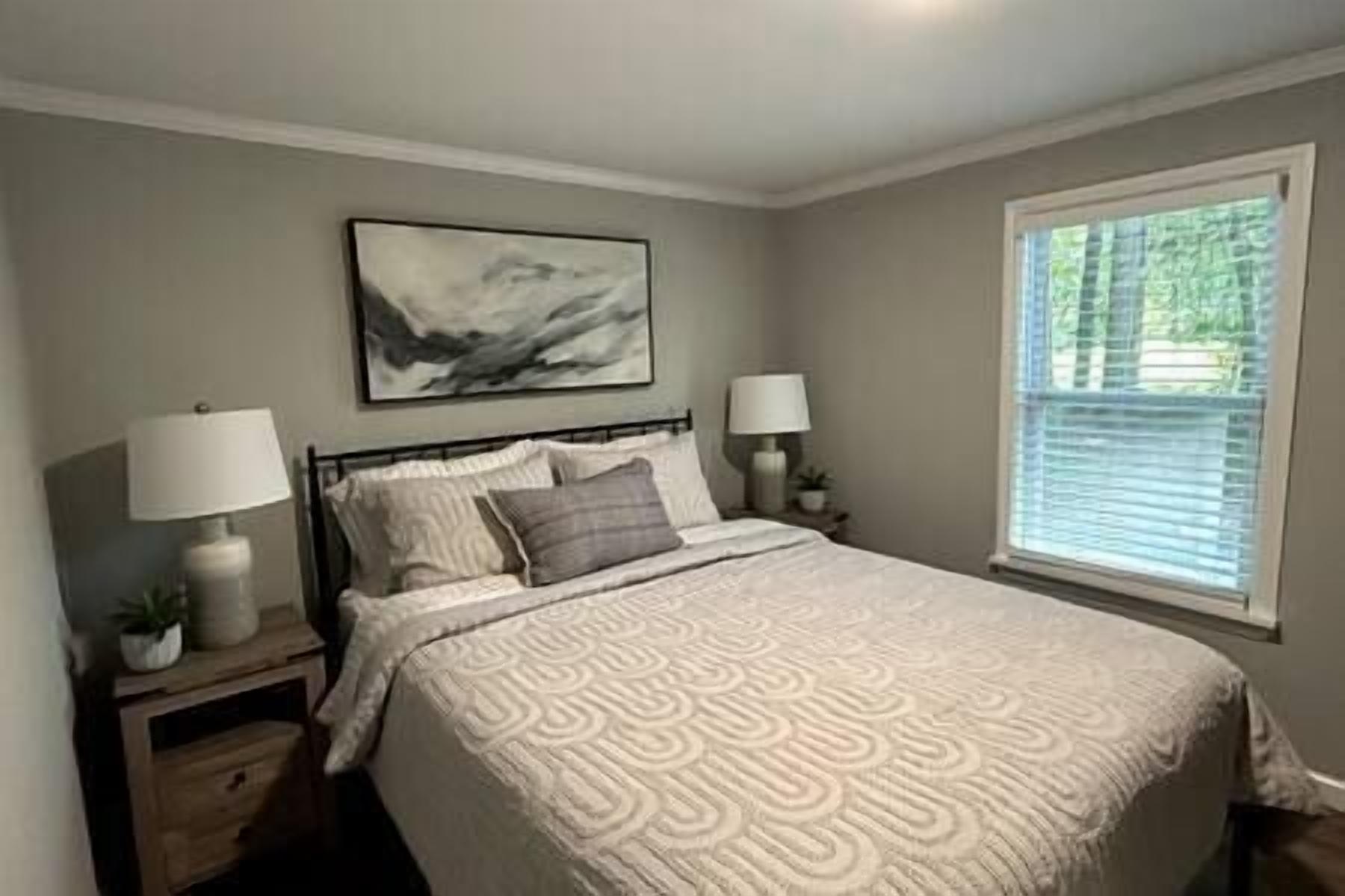 A bedroom with a bed, two bedside tables with lamps, a nightstand, a window with blinds, and a painting on the wall.
