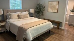 Een slaapkamer met een groot bed, nachtkastjes, een raam met zonwering, een plant en een schilderij aan de muur.