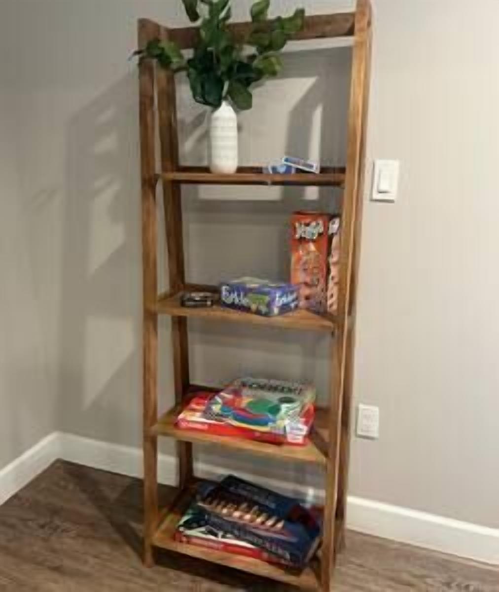 A wooden bookshelf with three shelves, a potted plant on the top shelf, and various items including a box of cereal and a board game.