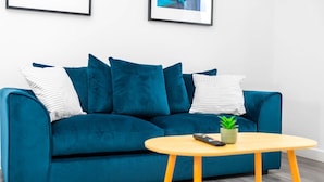 Ein blaugrünes Sofa mit gestreiften Kissen, ein kleiner runder Holztisch mit Fernbedienung und Pflanze sowie ein gerahmtes Bild an der Wand.