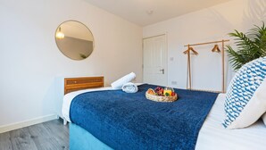 Une chambre à coucher avec un lit, un miroir rond, une tête de lit en bois, une couverture bleue, un panier de fruits et une plante suspendue.