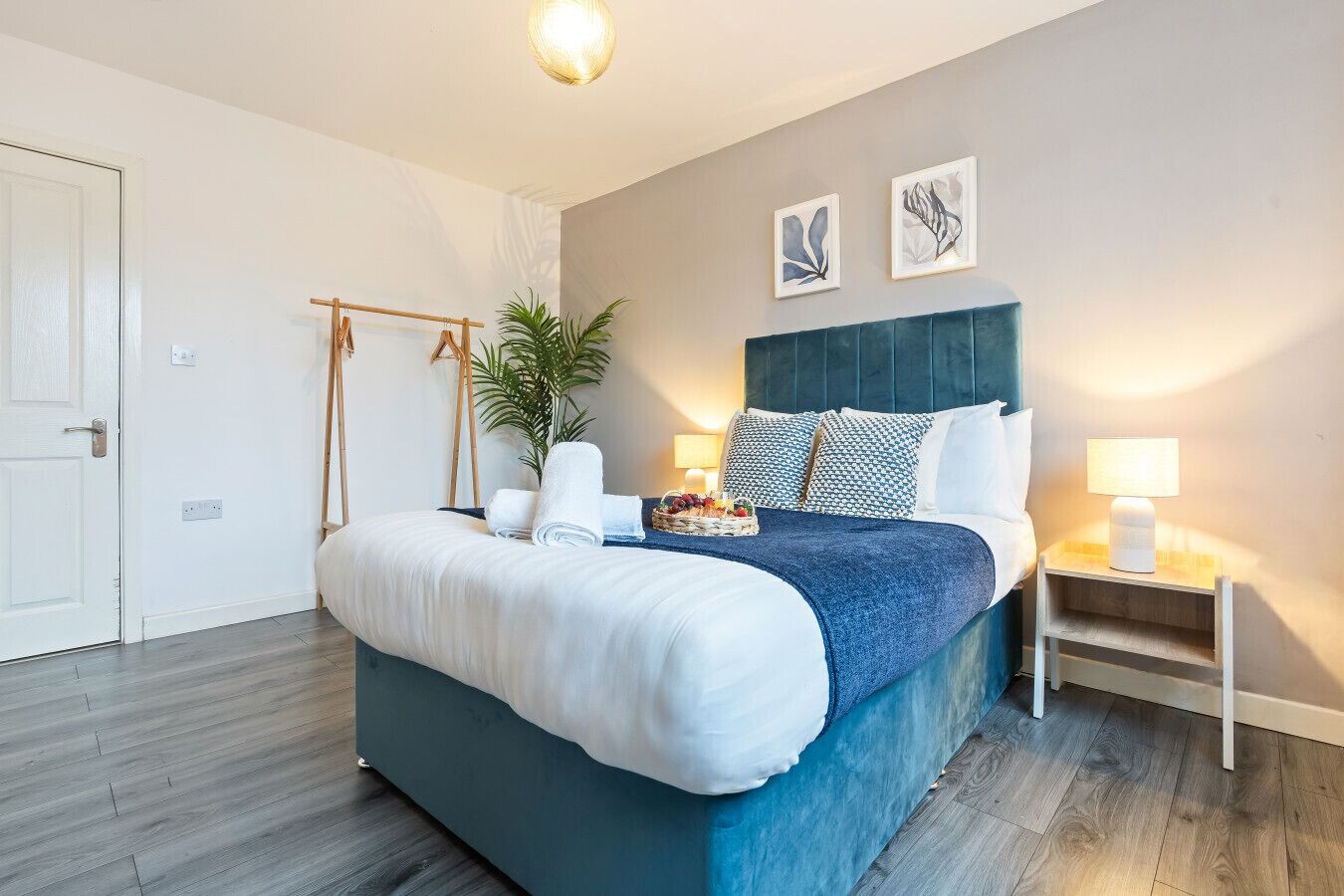 A modern bedroom with a large bed, a bedside table with a lamp, a wall-mounted shelf, and a potted plant.