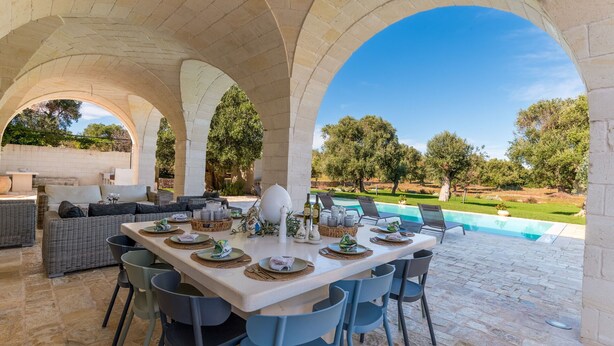An outdoor dining area with a pool view, stone arches, and outdoor furniture.