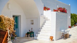 A white building with a staircase leading to a door, surrounded by potted plants and outdoor furniture.