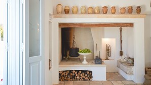 A rustic kitchen with a stone fireplace, a shelf with pottery, a bowl of apples, and a hanging brass instrument.