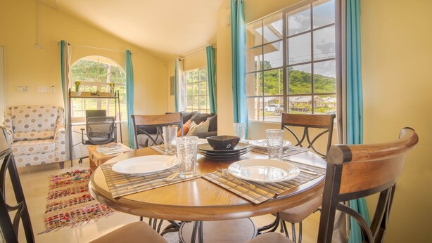 A dining area with a round table set for four, surrounded by chairs. There is a sofa and a chair in the background, and a window with curtains. - Casia Villa (Montego Bay)