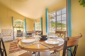 A dining area with a round table set for four, surrounded by chairs. There is a sofa and a chair in the background, and a window with curtains. - Casia Villa (Montego Bay)