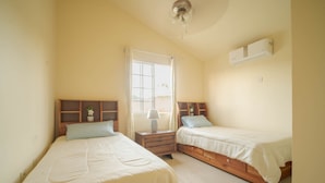 A bedroom with two single beds, a nightstand with a lamp, a window with curtains, and an air conditioning unit on the wall. - Casia Villa (Montego Bay)