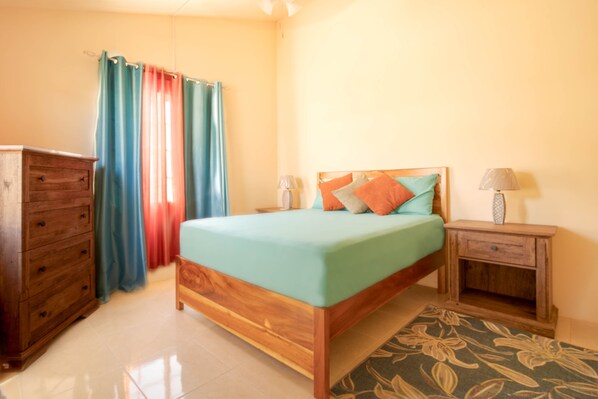 A bedroom with a wooden bed, a wooden dresser, a nightstand with a lamp, and a window with curtains. - Casia Villa (Montego Bay)