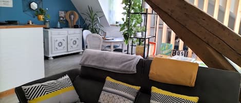 Un salon moderne avec un canapé en cuir noir, des coussins à motifs, un escalier en bois et, au fond, un coin repas.