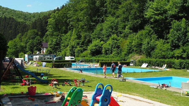 Un camping con piscina, zona de juegos infantiles y un paisaje montañoso cubierto de bosque al fondo.