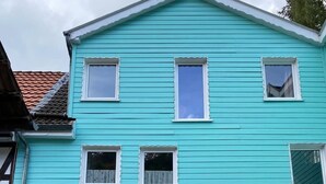 Un edificio de dos pisos con fachada color turquesa, varias ventanas, una puerta principal y un pequeño porche con plantas en macetas.