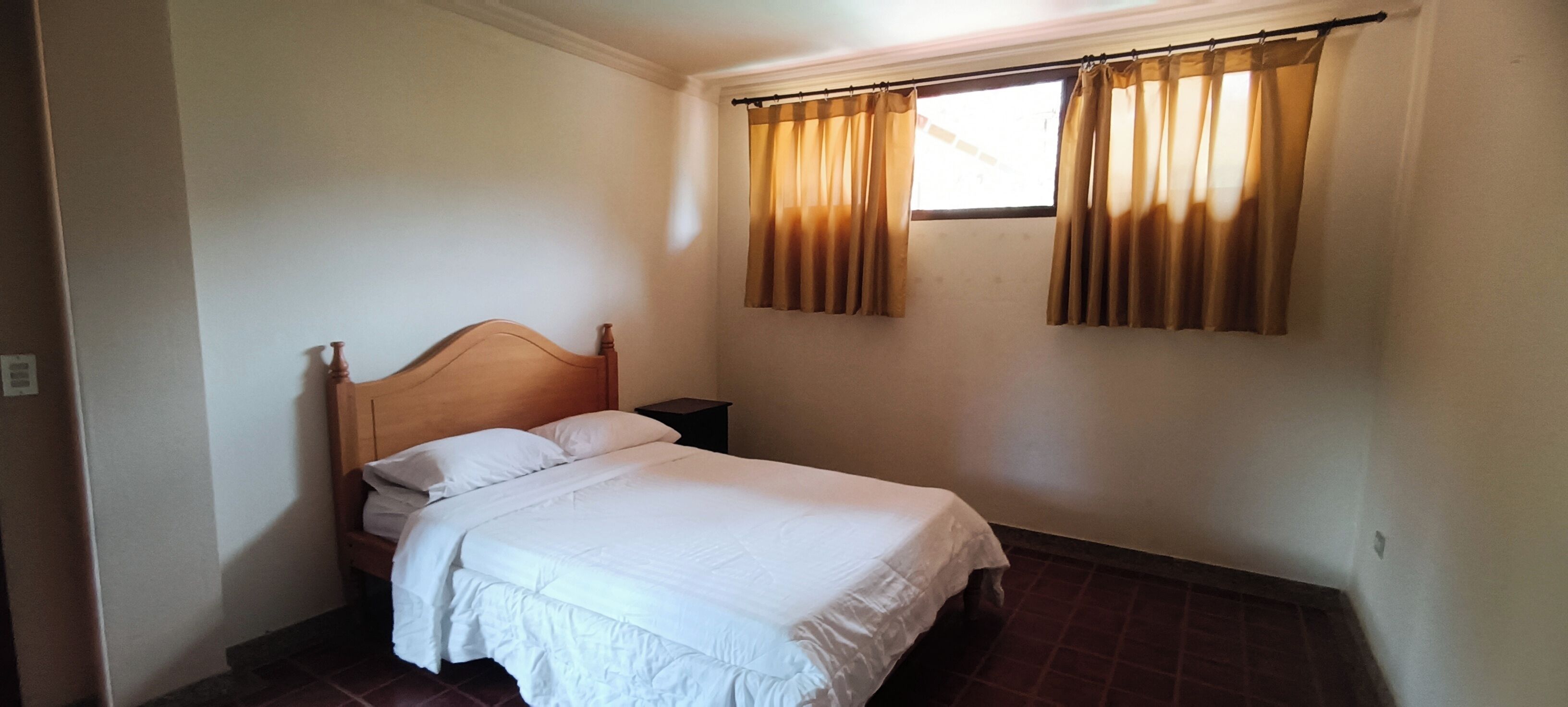 A bedroom with a wooden bed, white bedding, a nightstand, and a window with curtains.