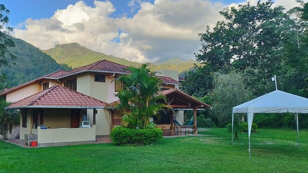 A two-story house with a red-tiled roof, surrounded by lush greenery and hills in the background.