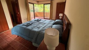A bedroom with a bed, a nightstand, a lamp, and a view of greenery through a sliding door.