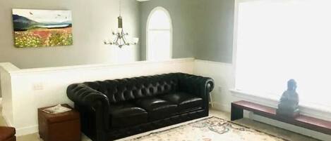Een woonkamer met een zwarte leren bank, een kamerkleed met patroon, een schilderij aan de muur en een kroonluchter.