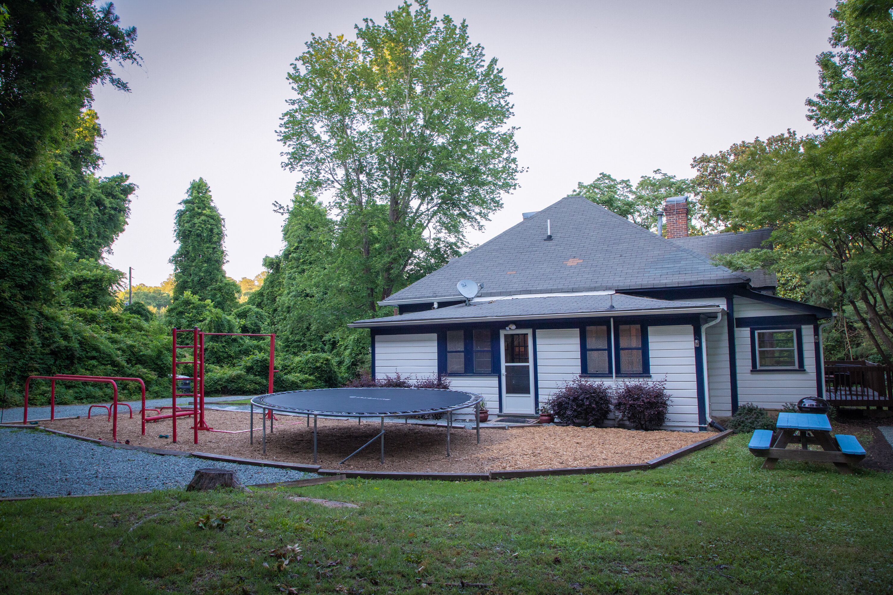 Una casa con patio trasero que incluye un trampolín, un módulo de juegos rojo y una mesa de picnic.