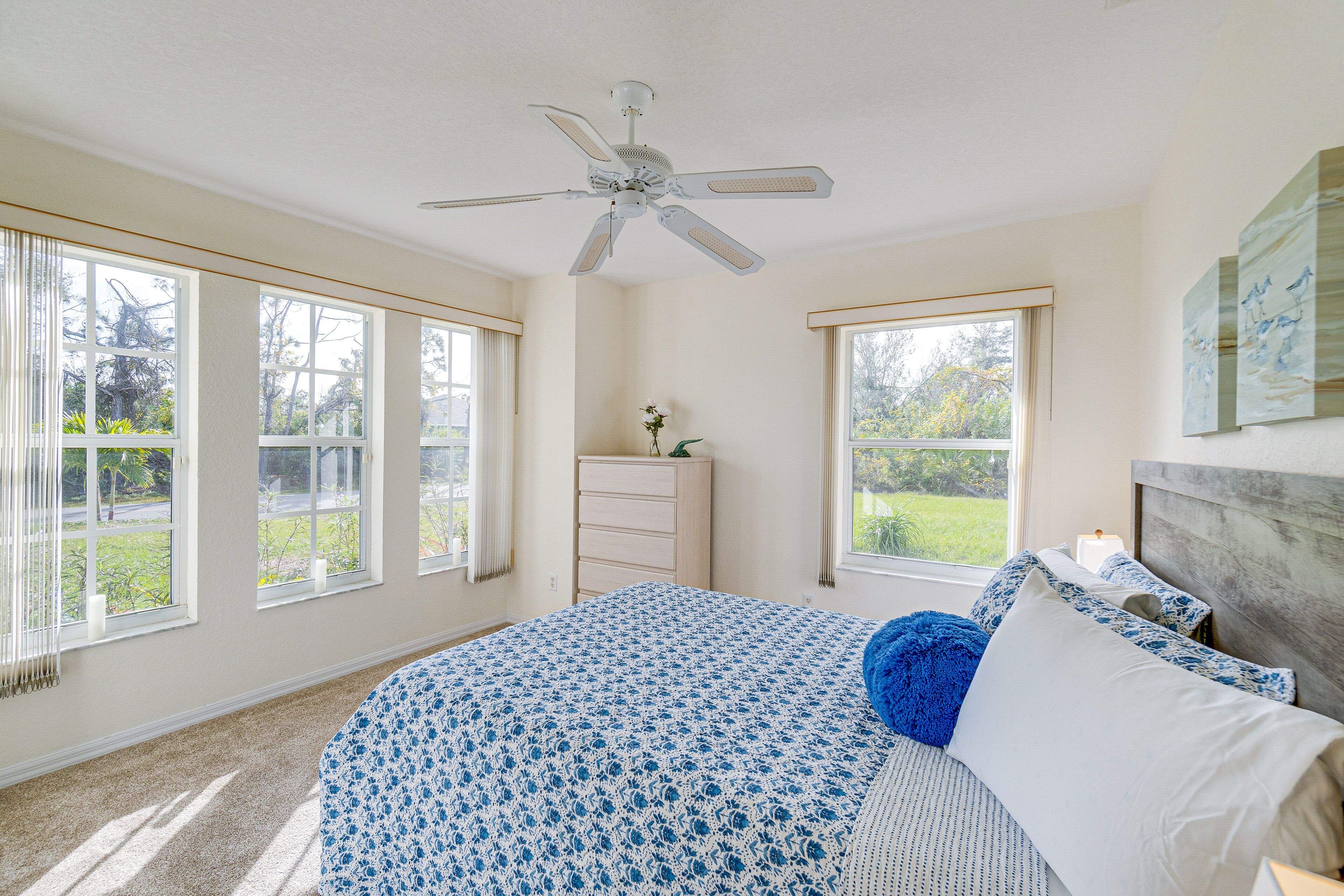 A bedroom with a bed, a dresser, a ceiling fan, and large windows.