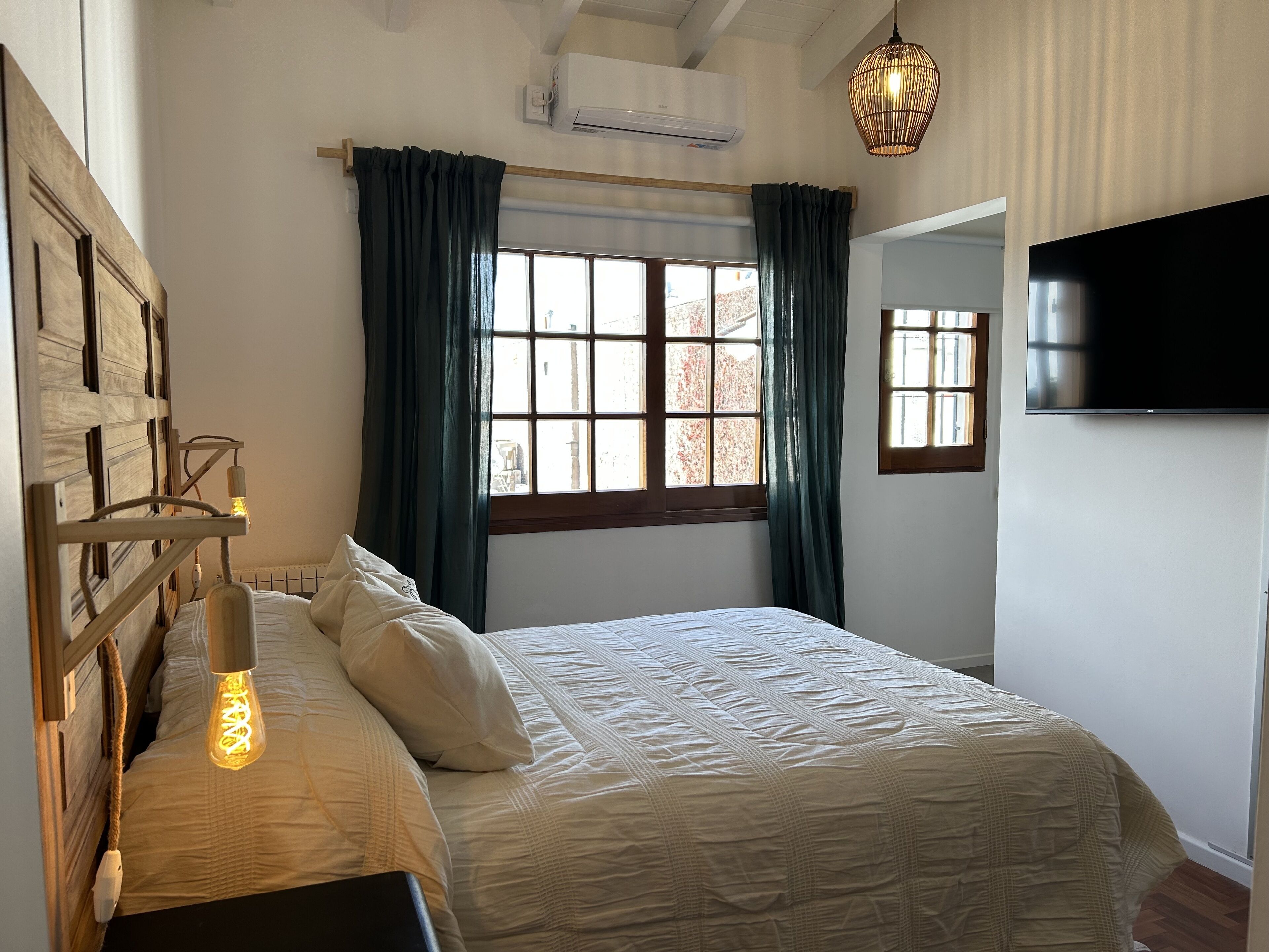 Une chambre à coucher comprenant un lit, une lampe de chevet, une fenêtre avec des rideaux, un appareil de climatisation et un téléviseur fixé au mur.